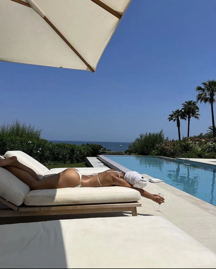 dddc395afb84fe56452809742942b42d_048cc0a5-95b3-4a1a-9da2-930296d143f5 A woman in a bikini relaxes on a lounge chair beside a sparkling pool under a clear blue sky.