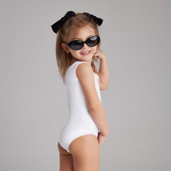 Young girl in a white swimsuit and black sunglasses smiles while posing playfully, wearing a black bow in her hair against a gray backdrop.