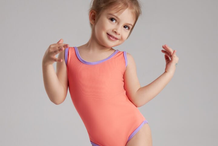 A little girl in a pink bodysuit smiles and poses playfully for the camera.