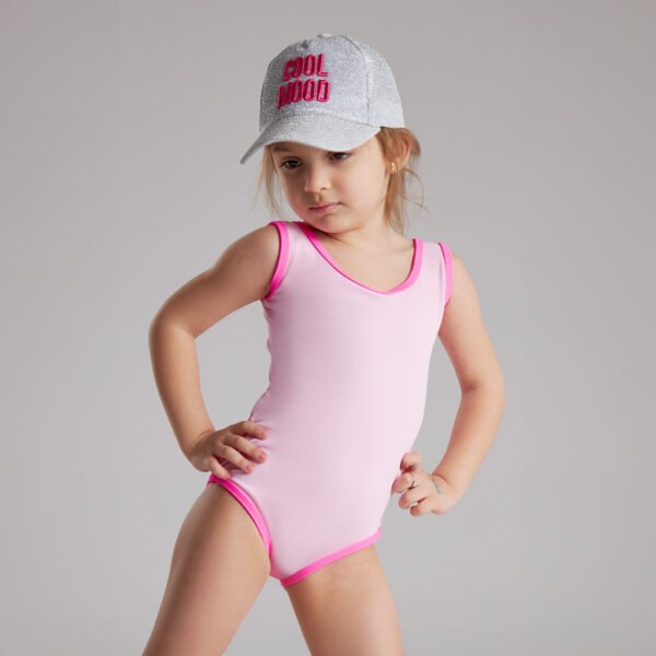 A little girl wearing a pink swimsuit and hat smiles while playing by the water.