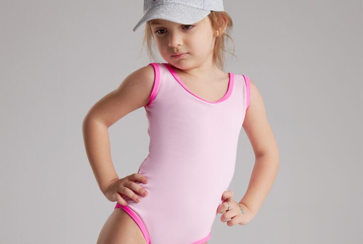 A little girl wearing a pink swimsuit and hat smiles while playing by the water.