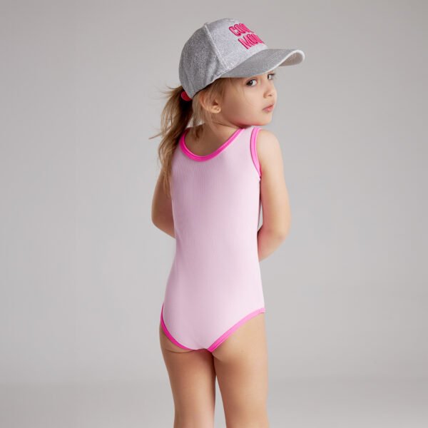 A little girl in a pink swimsuit and a baseball cap smiles playfully at the camera.