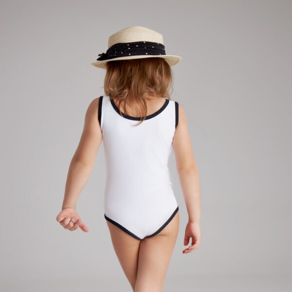Back view of young girl wearing a white one-piece swimsuit with black trim and a straw hat, walking confidently indoors.