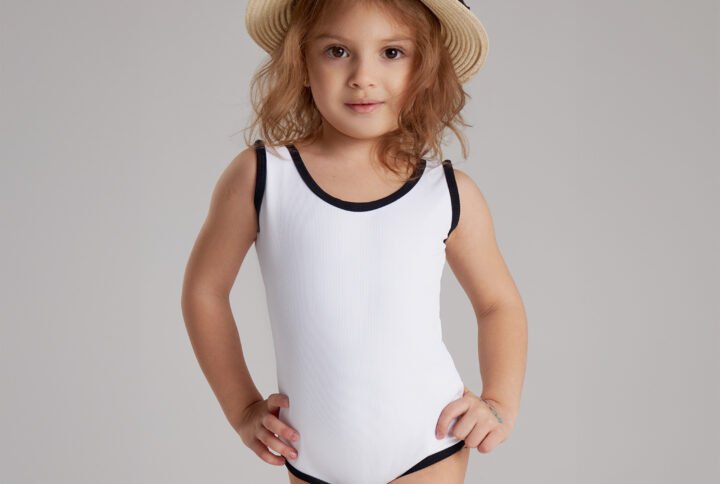 Young girl wearing a white one-piece swimsuit with black trim and a straw hat, posing confidently in studio setting.